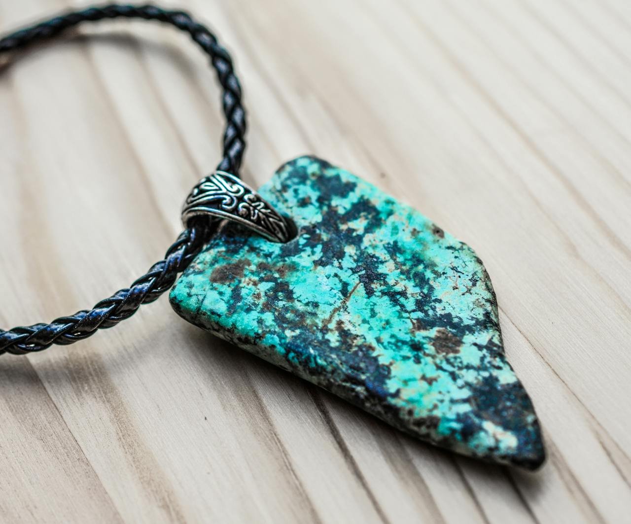 A detailed close-up of a turquoise arrowhead pendant with a braided leather chain on a wooden surface.